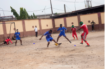 A group of visually impaired players engaged in a soccer match on a dusty, uneven field enclosed by a high wall. Two teams, one wearing blue jerseys and the other in red, are actively competing for the ball. The players are using a specially designed ball with sound to navigate the game. One player in blue is attempting to dribble past an opponent in red, while other players, including a goalkeeper near a simple goalpost, are positioned around the field. Some players are wearing blindfolds, and a few spectators stand in the background watching the game.