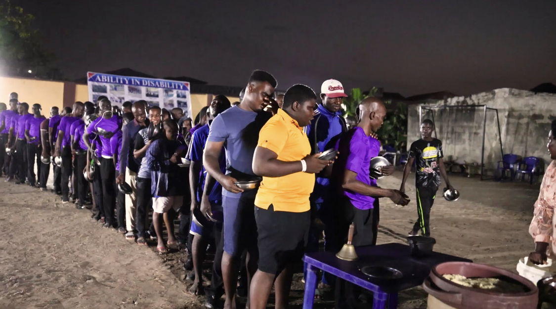 A long line of people, mostly wearing purple shirts, waiting outdoors in the evening to receive food. Some individuals are holding plates or bowls while standing in line. A man in a yellow shirt is at the front, looking at his phone. In the background, there is a banner with text and additional people standing near the serving area.