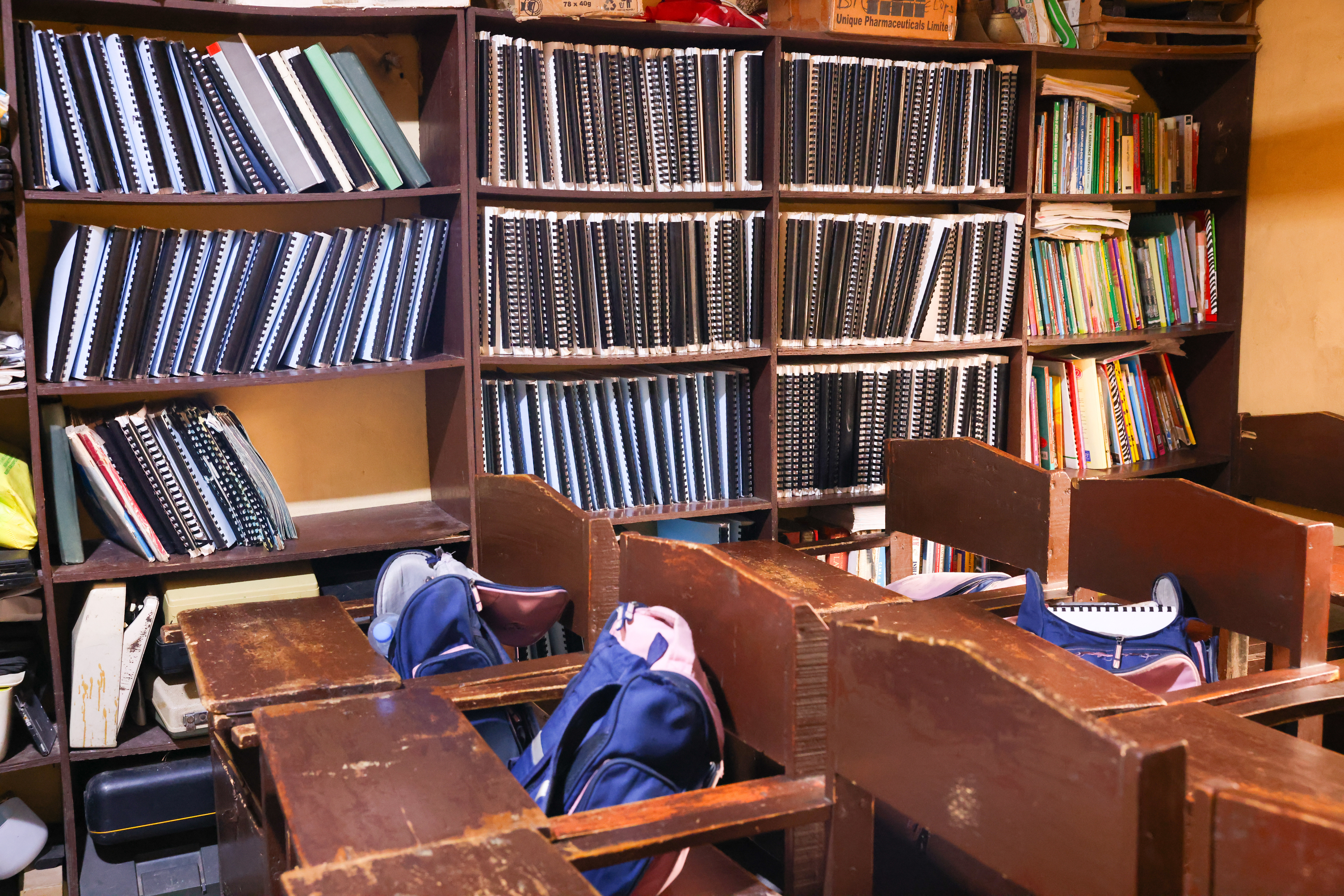 A classroom with wooden desks and chairs, each with a school bag placed on it. The background features large bookshelves filled with neatly arranged black spiral-bound books, along with some colorful textbooks and other materials. The room has a warm, slightly aged atmosphere.