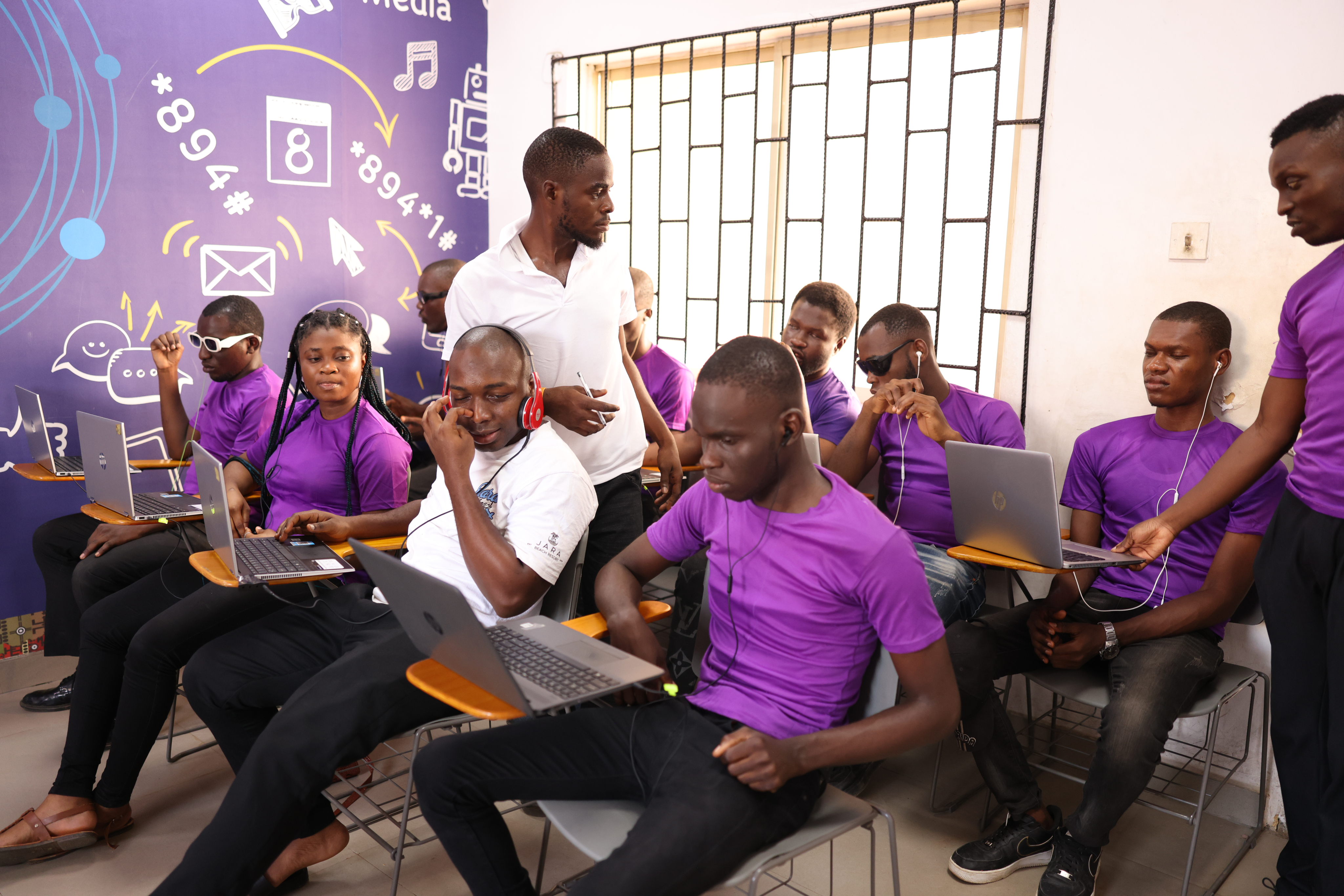 A group of young people wearing matching purple shirts sitting in a classroom, each using a laptop and wearing earphones. A man in a white shirt is standing and engaging with them, possibly instructing or assisting. The background features a wall decorated with technology and social media icons, adding a digital learning atmosphere.