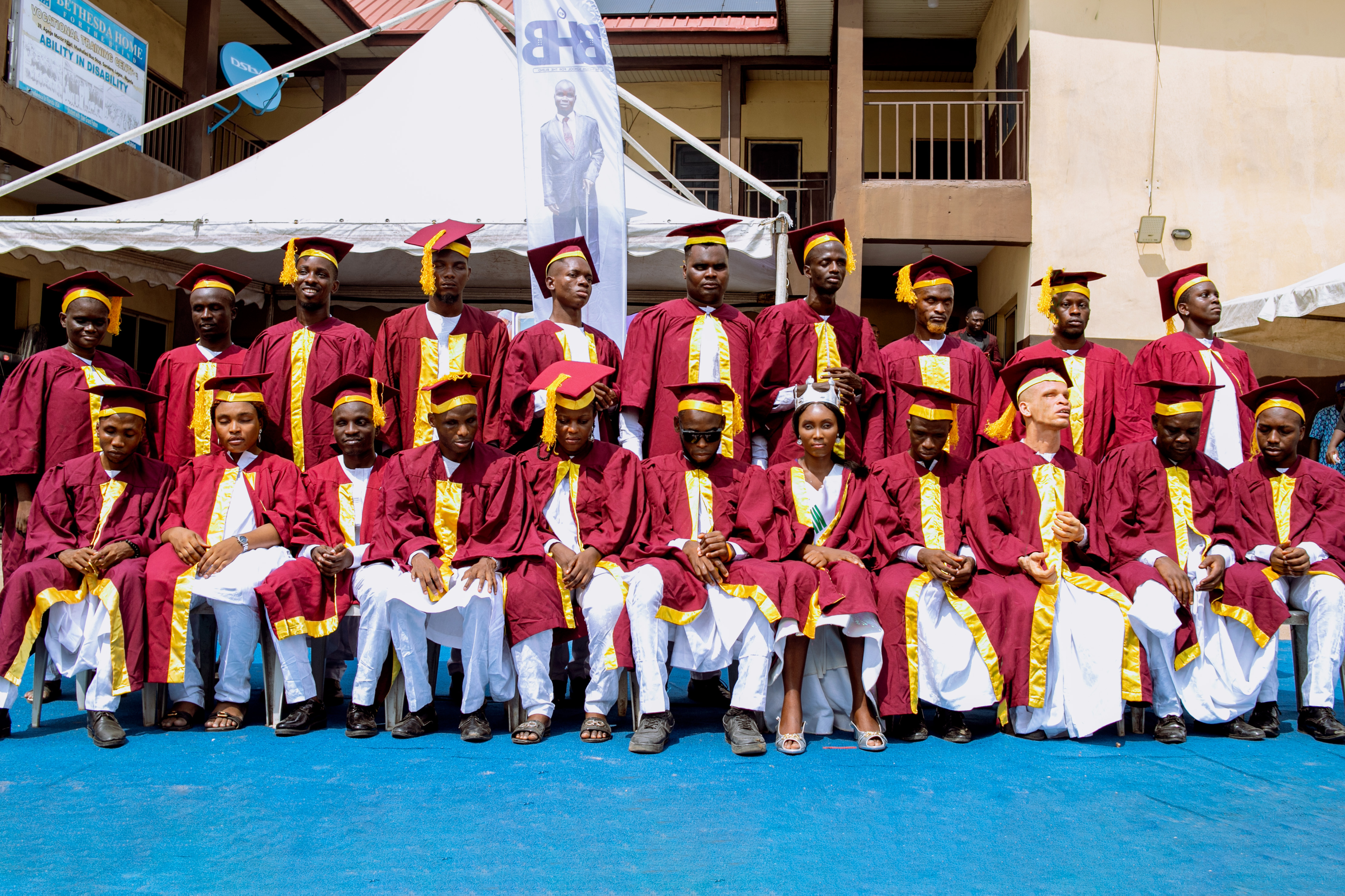 A group of visually impaired graduates dressed in maroon and gold academic gowns and caps, seated and standing in rows on a blue stage at an outdoor graduation ceremony. The background features a white tent, a banner with a man's image, and a building with balconies. The graduates have various expressions, some smiling and others serious.