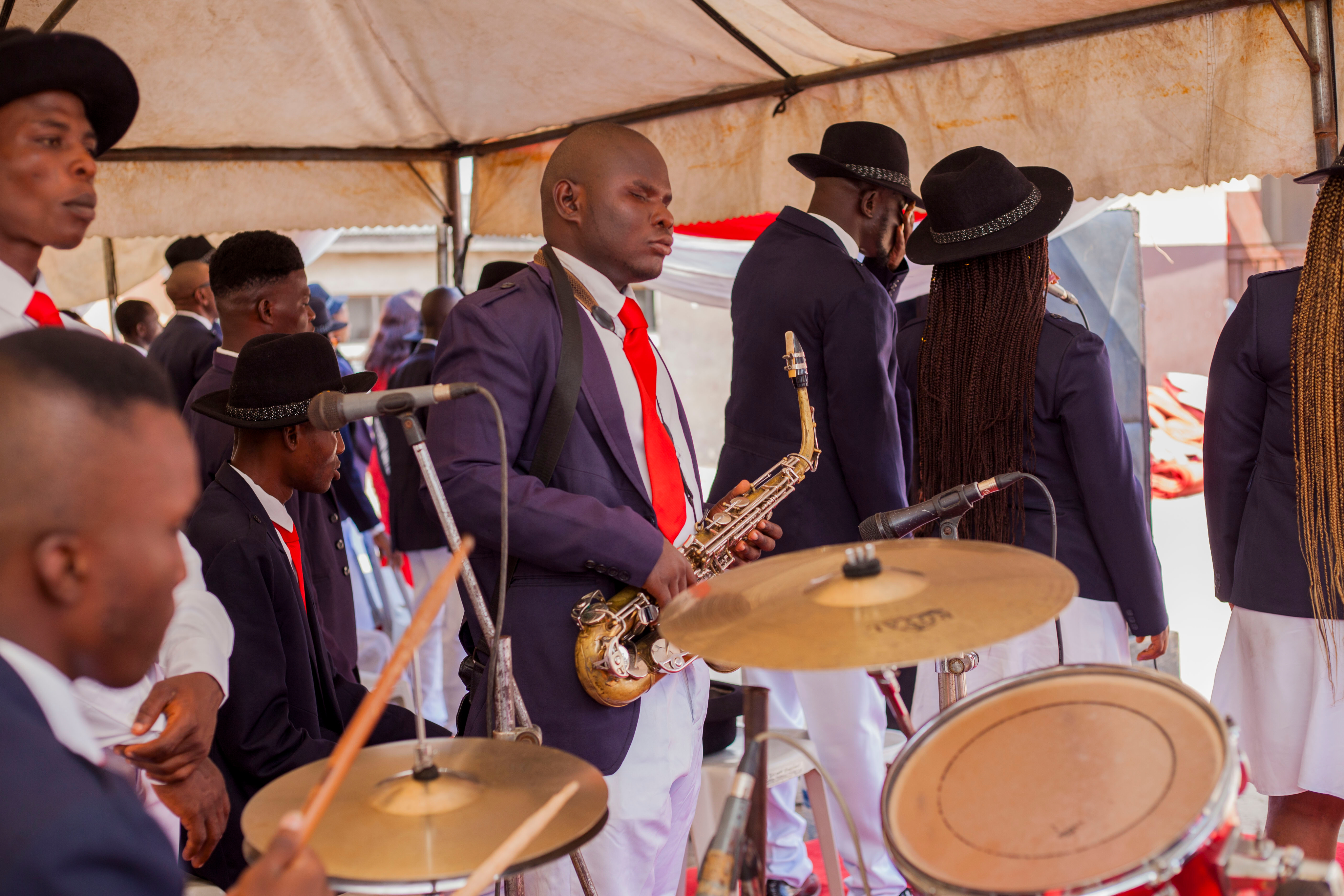 A group of visually impaired musicians performing under a tent. They are dressed in navy blue suits with white shirts and red ties. A man in the foreground is holding a saxophone, while others are playing drums and other instruments. Some musicians are wearing black hats. The atmosphere suggests a formal event or celebration, with the performers focused on their music