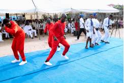 Visually impaired dancers performing in a dance competition on a blue stage. Some are dressed in red outfits while others wear white, moving energetically in sync. A crowd of spectators watches from under a tent in the background, indicating a formal event or festival.