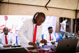 An image of a visually impaired DJ wearing a white shirt and red tie, mixing music on a DJ controller under a tent. He is wearing headphones and focused on his equipment.